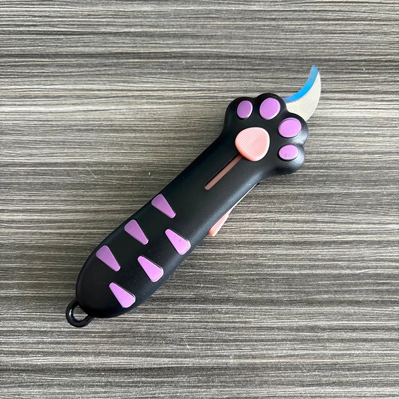 A black utility knife shaped like a cat paw with purple accents and a light blue blade resting on a gray wooden surface.
