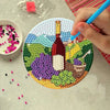A hand uses a blue tool to place a gold-colored bottle design onto a mosaic coaster featuring various colored beads and a lan