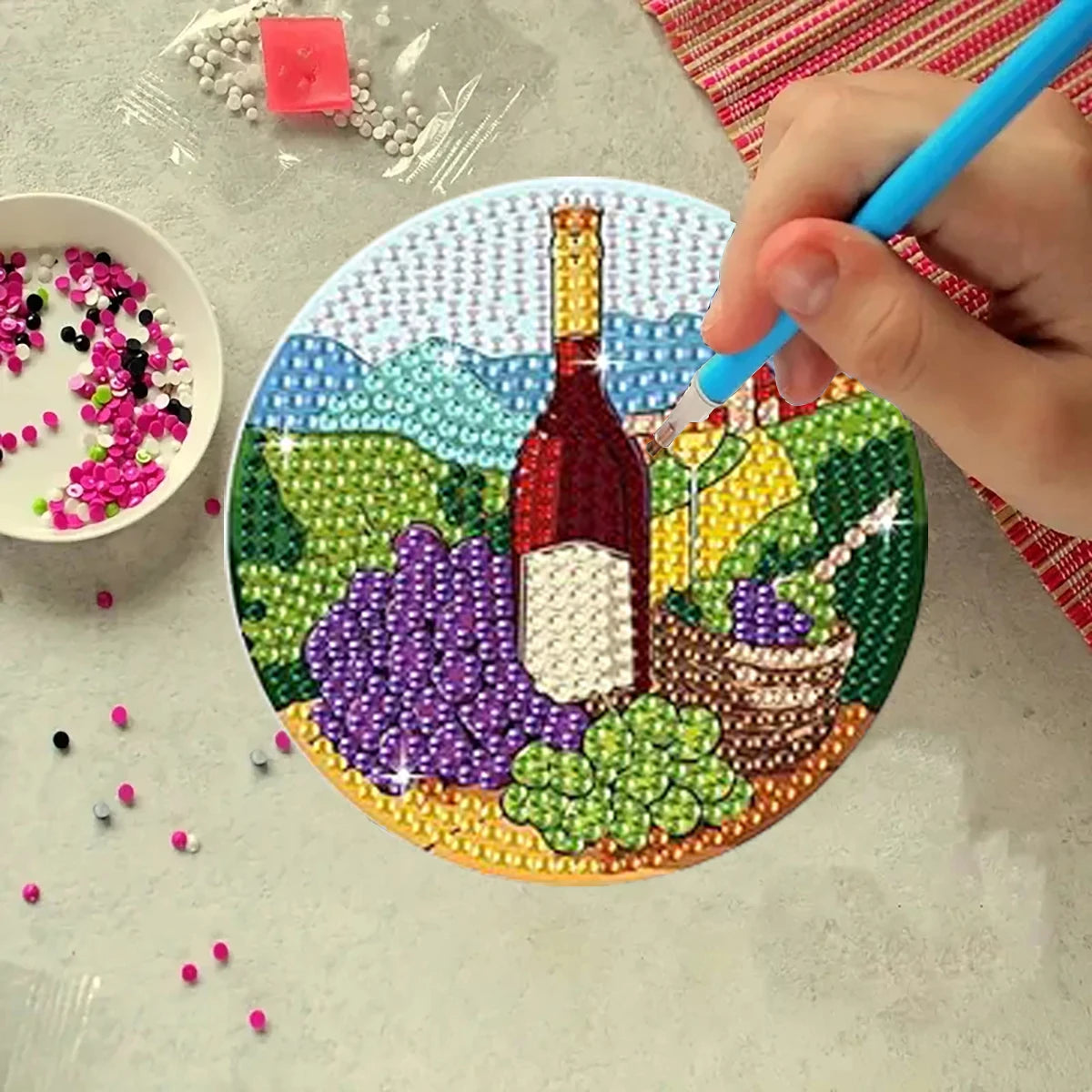 A hand uses a blue tool to place a gold-colored bottle design onto a mosaic coaster featuring various colored beads and a lan