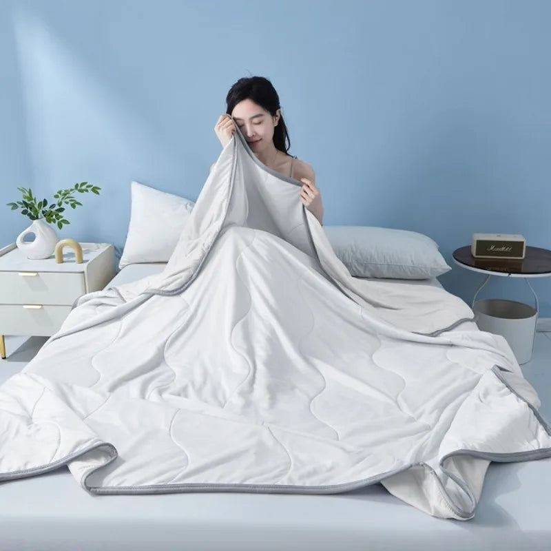 A white cooling summer quilt is draped on a bed, with a woman resting her head on the blanket in a light blue room.