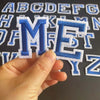 A hand holds an embroidered blue and white alphabet patch letter ME from a set of iron-on letters.