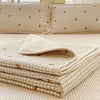 A stack of beige floral quilted bedspreads with gold accents resting on a light yellow surface.