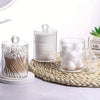 Clear acrylic jar filled with white cotton balls and swabs on a woven mat.