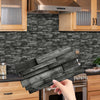 A hand holds a bundle of dark wood tile stickers against a gray stacked brick backsplash in a kitchen setting.