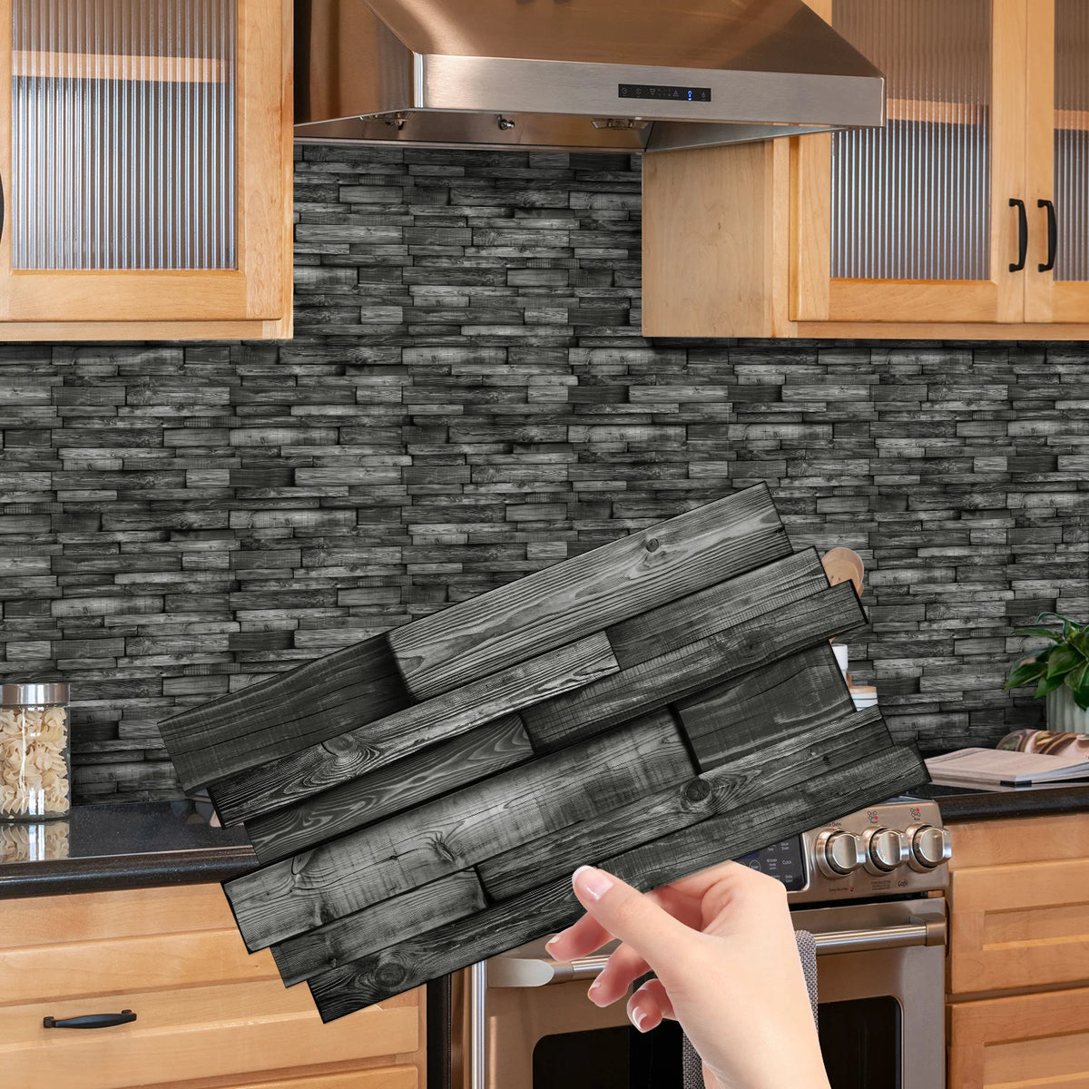 A hand holds a bundle of dark wood tile stickers against a gray stacked brick backsplash in a kitchen setting.