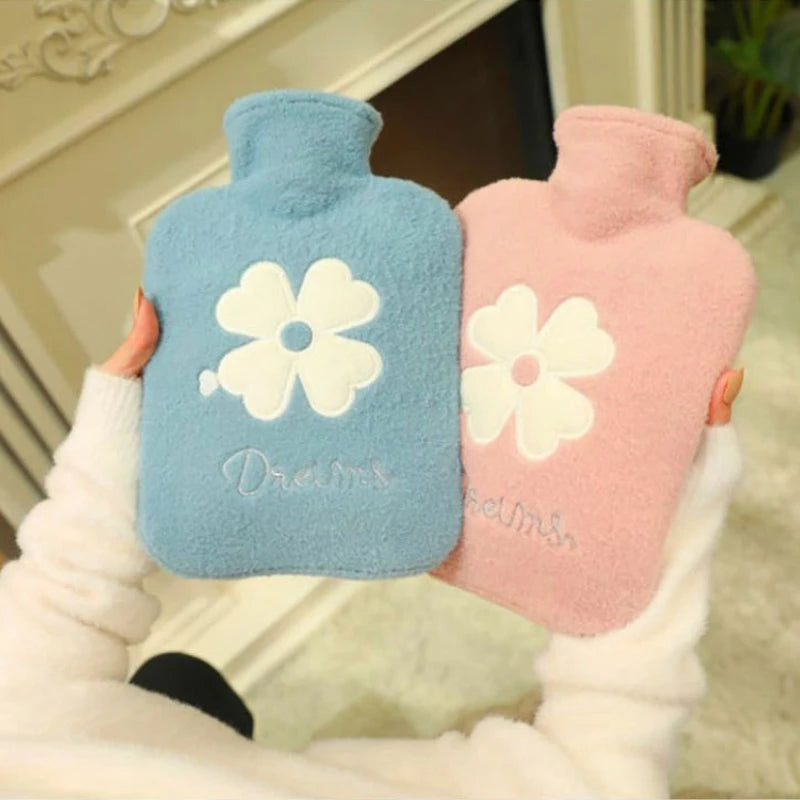 A hand holds a light blue plush-covered hot water bottle with a white flower design and a pink plush-covered hot water bottle