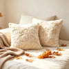 A collection of cream-colored plush throw pillows featuring embroidered autumn pumpkin and maple leaf designs on a white bedd