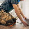 A person wearing a dark shirt and work pants is using heavy-duty knee pads while working on a wooden surface.