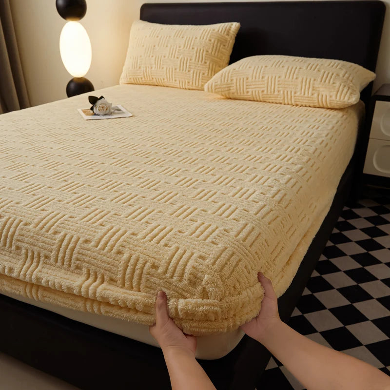 A beige plush mattress cover is displayed on a black bed, with a yellow lamp and a small item resting on the cover.