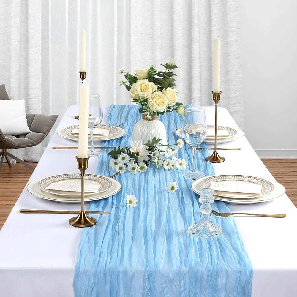 A white table setting featuring a blue sheer gauze table runner, gold candlesticks with white candles, and floral arrangement