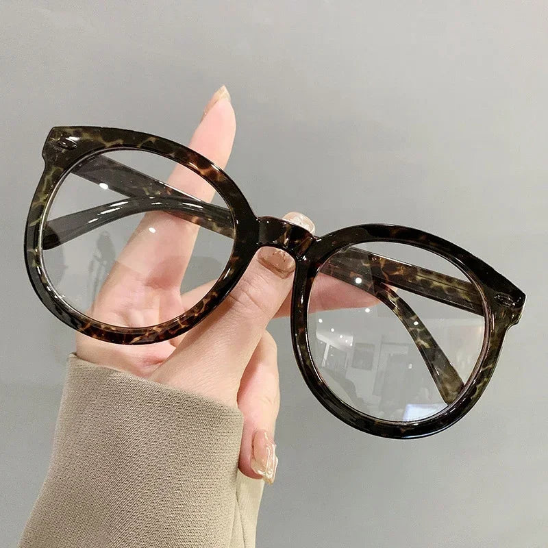 Eine Hand hält eine große braune Schildpatt-Brille mit klaren Linsen vor einem hellgrauen Hintergrund.