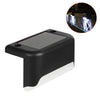 A black solar deck light with a clear lens illuminates a wooden railing outdoors.