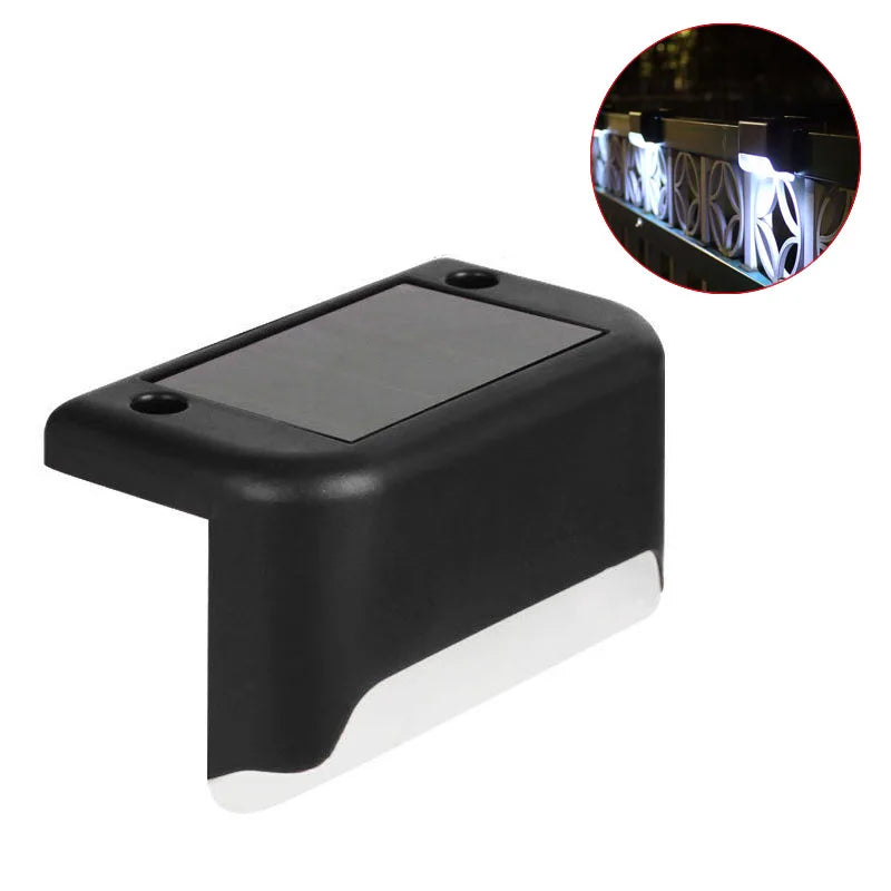 A black solar deck light with a clear lens illuminates a wooden railing outdoors.