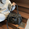 A black soft leather plaid crossbody bag with a long strap is shown resting on a wooden shelf.