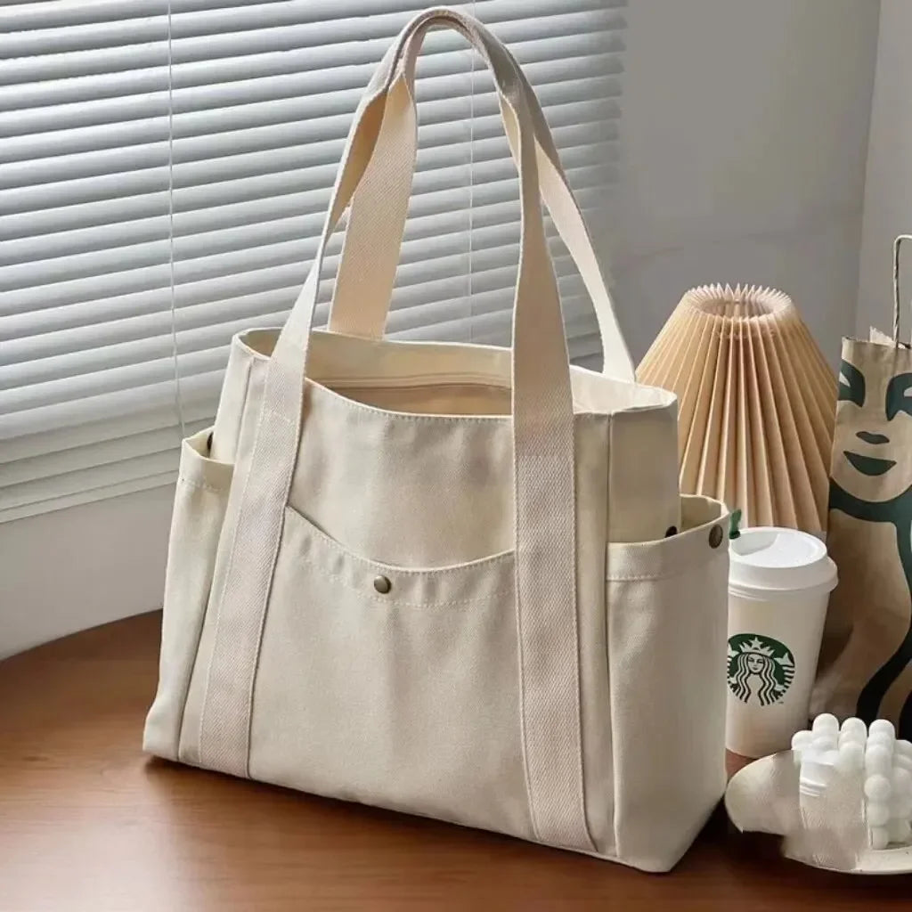 Una bolsa de lona beige con tirantes color canela, mostrada sobre una superficie de madera junto a una planta en maceta.