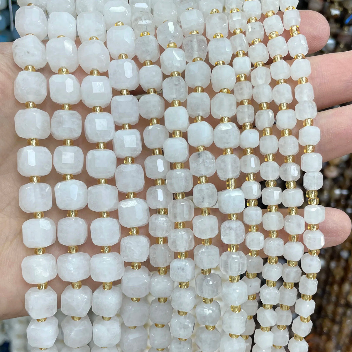 A hand holds a strand of square faceted natural stone beads in white and translucent tones for jewelry making.