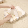 Hands are placing rolled fabric items into a clear packing cube set on a light beige textured surface.