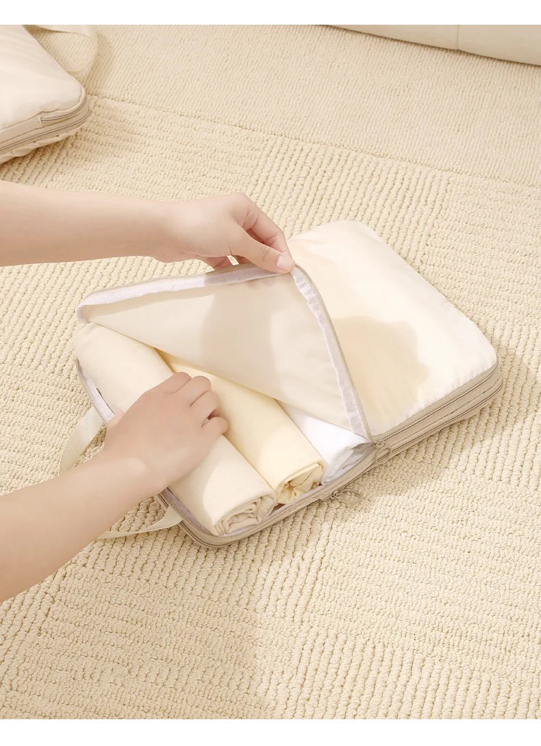 Hands are placing rolled fabric items into a clear packing cube set on a light beige textured surface.