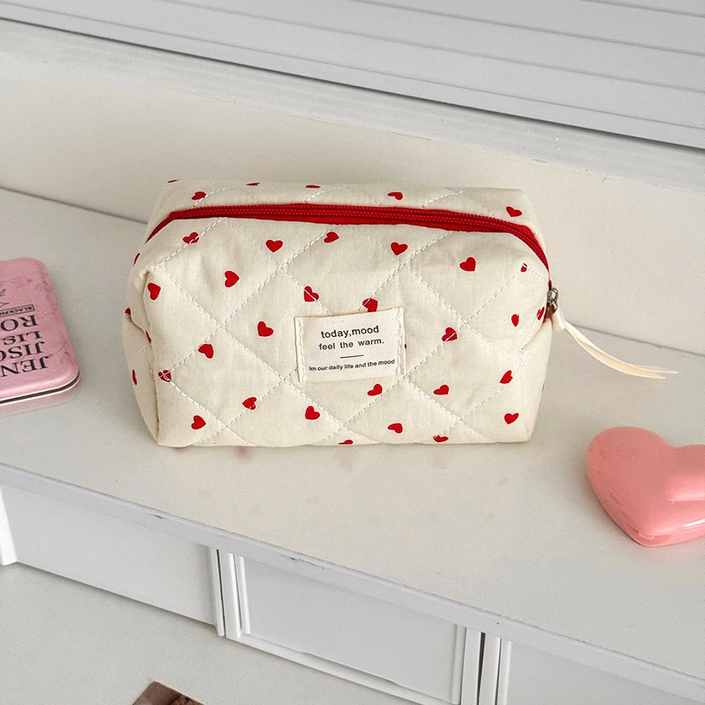 A quilted white makeup bag with red heart embroidery and a red zipper lying on a white shelf.