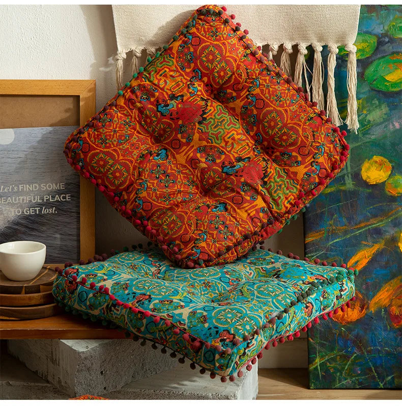 A vibrant orange and red bohemian cotton linen floor cushion with fringe hanging against a colorful painting.