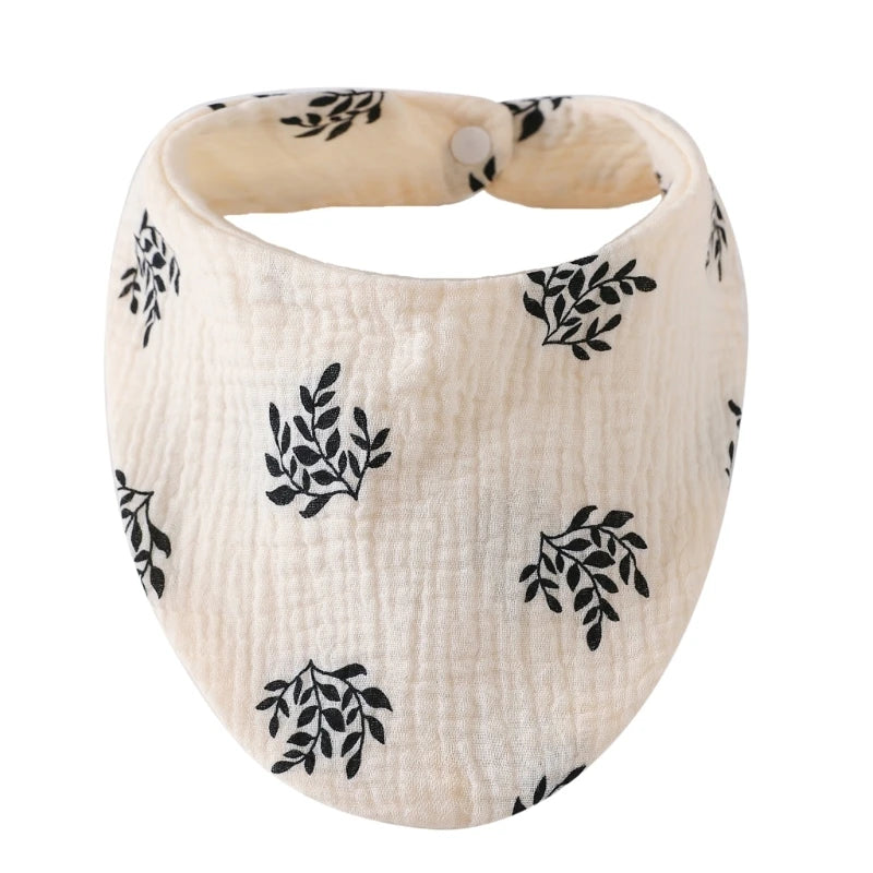 A beige organic cotton muslin baby bib with a gathered band for absorbent use, featuring a black leafy print.