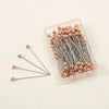 A clear plastic container holds a pack of 100 precision quilting pins with rose gold colored heads and silver shafts.