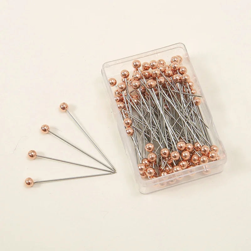 A clear plastic container holds a pack of 100 precision quilting pins with rose gold colored heads and silver shafts.