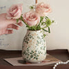 Eine Hand hält eine rosa Keramikvase, gefüllt mit hellrosa Rosen und weißen Füllblumen.