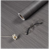 A roll of self-adhesive wood grain wallpaper film next to a pair of eyeglasses and a small flower on a gray textured surface.