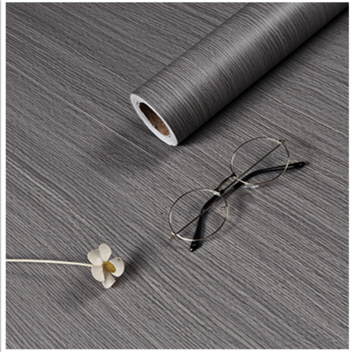 A roll of self-adhesive wood grain wallpaper film next to a pair of eyeglasses and a small flower on a gray textured surface.