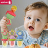 A baby is holding a green silicone fruit feeder with a dinosaur design, next to other colored feeders.