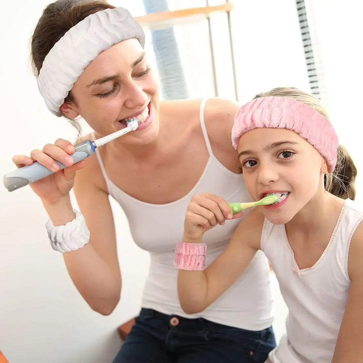 A woman is brushing a child's teeth while the child wears a pink headband and holds a toothbrush.