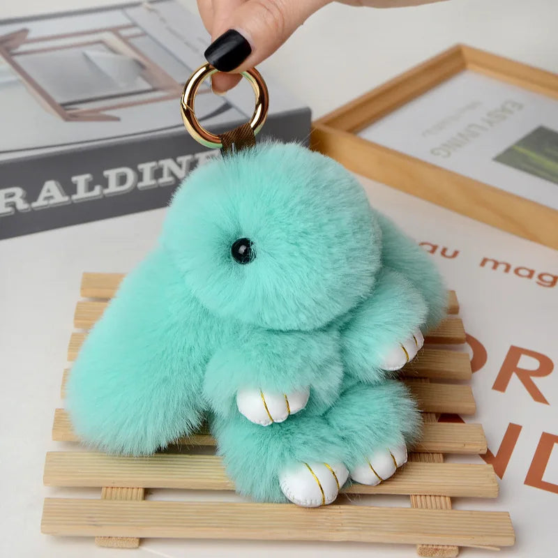 A fluffy turquoise bunny keychain with a pom pom tail resting on a wooden surface.