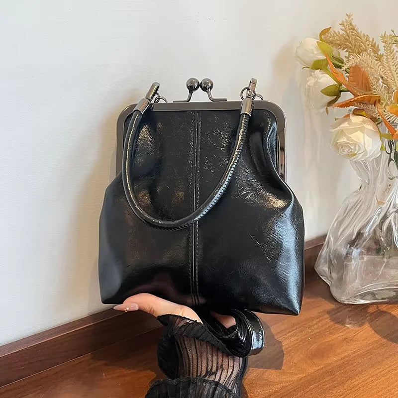 A black leather crossbody handbag is displayed against a white wall next to a vase of white roses.