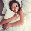 A smiling woman is resting her head on a white pillow, embracing the bedding.