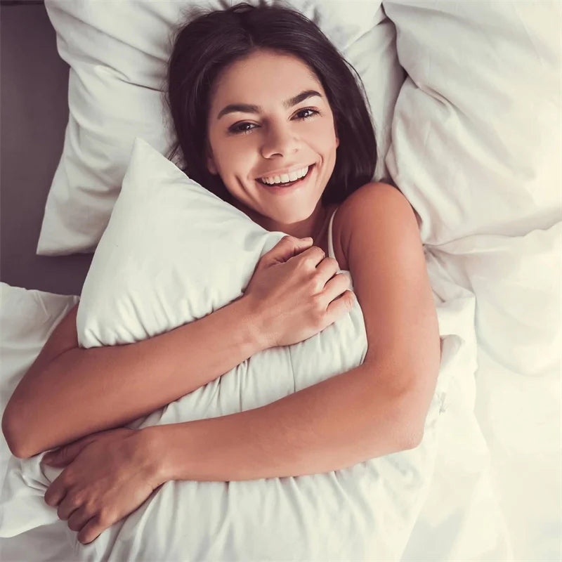 A smiling woman is resting her head on a white pillow, embracing the bedding.