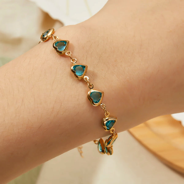 Nahaufnahme eines goldenen Armbandes mit kleinen, durchscheinenden blauen Kristallherzanhängern am Handgelenk.