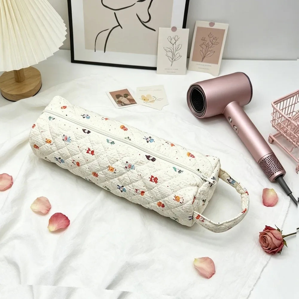 A pink quilted cotton hair dryer storage bag with a handle resting on a white cloth next to a framed picture.