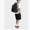 A young man wearing a tan shirt and black shorts carries an olive green laptop backpack.