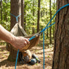 A hand is using a blue rope to tie or adjust a heavy-duty rope tensioner tool near a tree trunk in a forest setting.