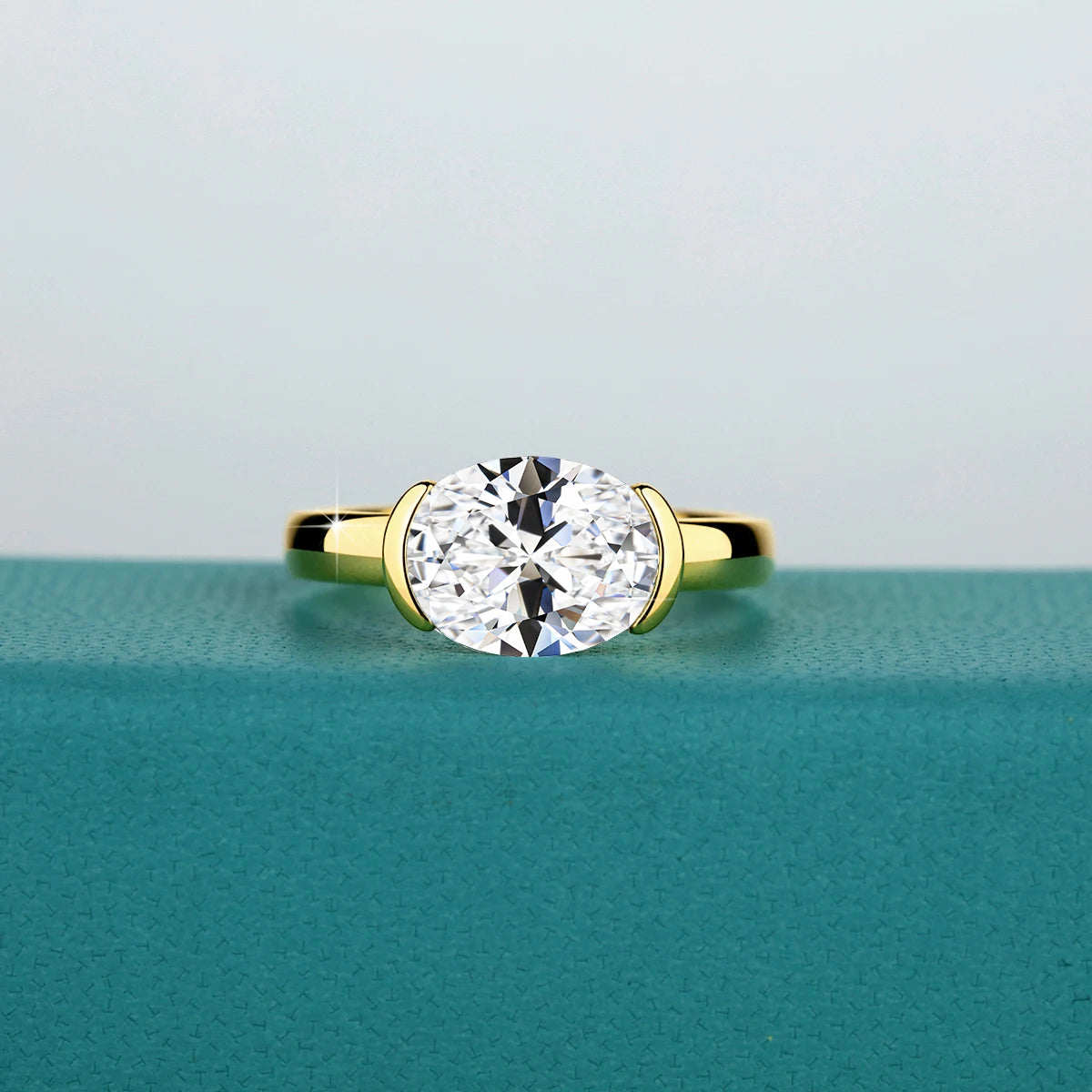 A close-up of a 14K gold oval moissanite ring with a brilliant clear stone resting on a teal surface.