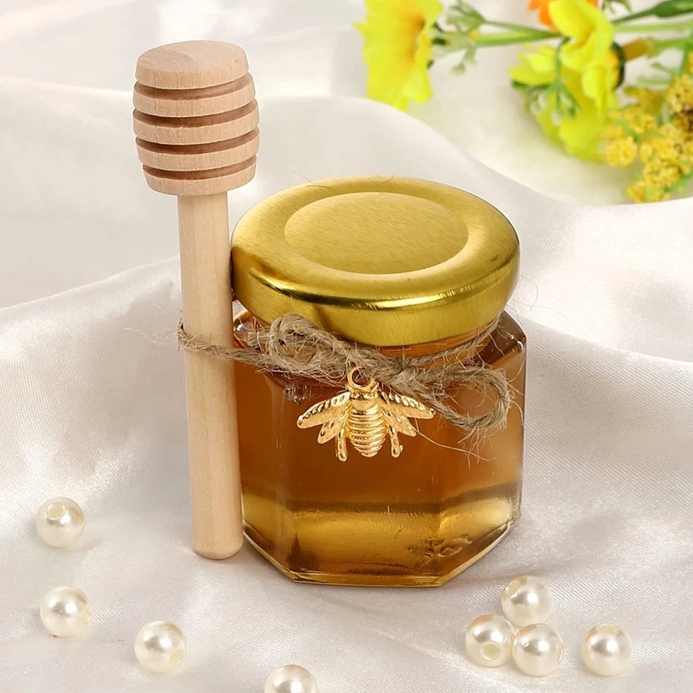 Mini glass honey jars with gold lids and wooden dippers displayed on white satin fabric.