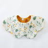 A white cotton baby feeding bib with a scalloped edge and embroidered green and yellow leaves, featuring a brown fabric backi