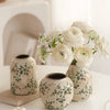 Weiße Keramikvase mit grünen Blumenmustern hält einen Strauß weißer Rosen und Babyblumen.