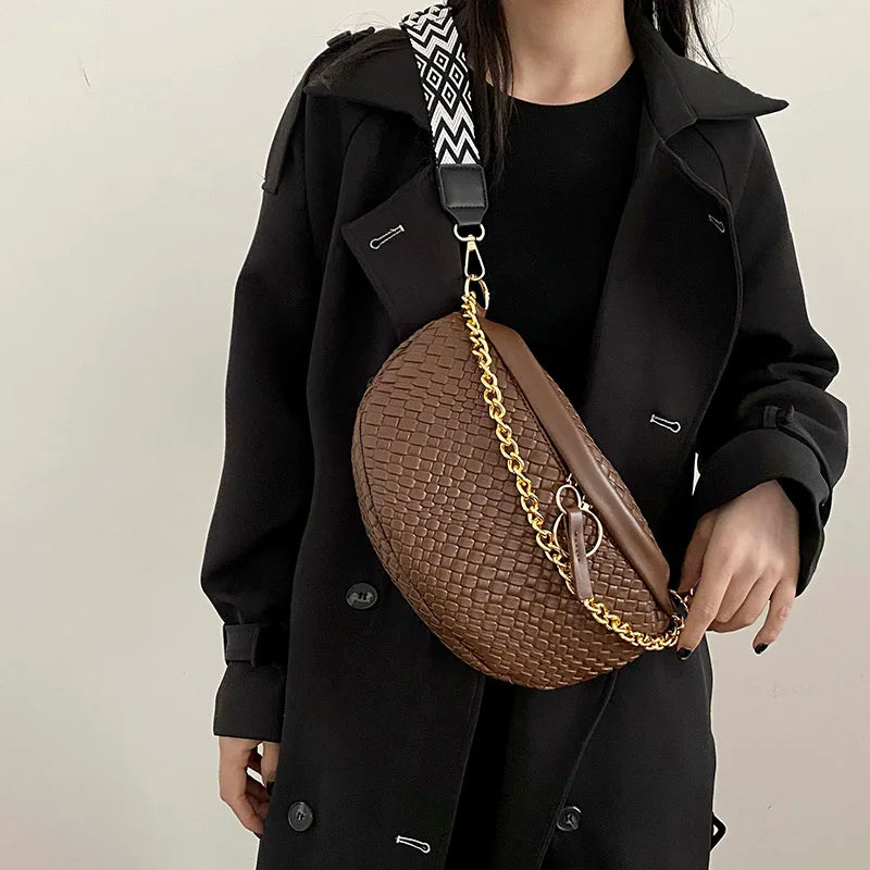 A woman wearing a black coat holds a brown woven pattern waist bag with a gold chain strap.