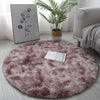 A round, shaggy mauve area rug placed on a light wooden floor next to a large potted plant and a sofa.