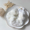 A white plush coil hair tie is displayed in a decorative white dish next to a natural woven sphere.
