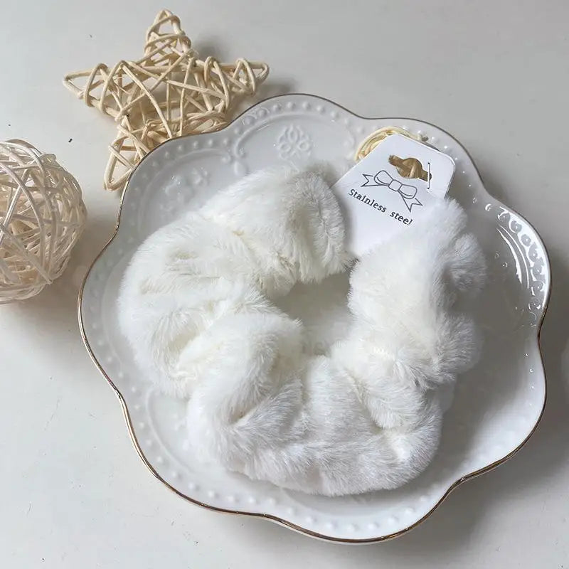 A white plush coil hair tie is displayed in a decorative white dish next to a natural woven sphere.