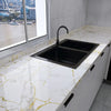 A modern kitchen sink installed in a white countertop with gold marble veining and black fixtures.