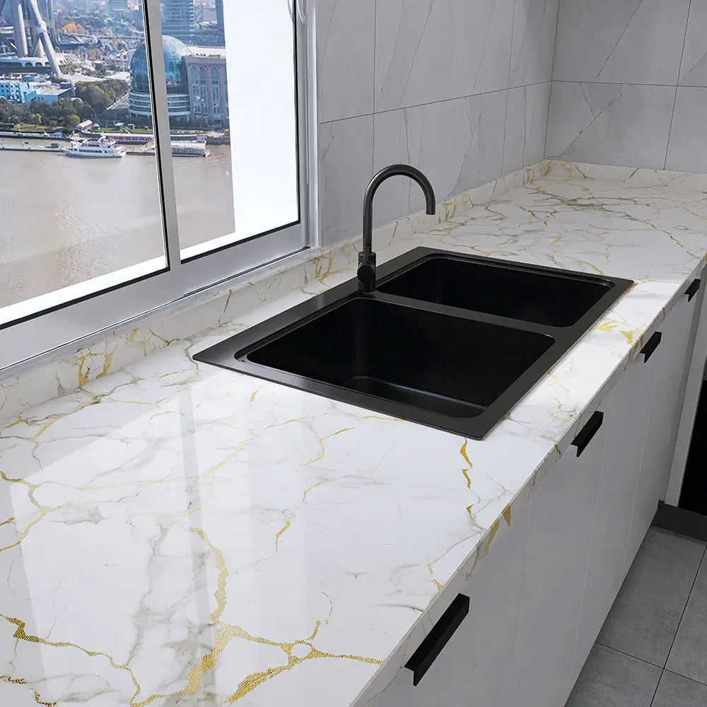 A modern kitchen sink installed in a white countertop with gold marble veining and black fixtures.